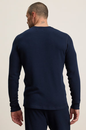 A man stands facing away, showcasing the back of the Velvet by Graham & Spencer TOBIAS CREW—a long-sleeve, dark blue thermal shirt—paired with matching pants and revealing a small neck tattoo.