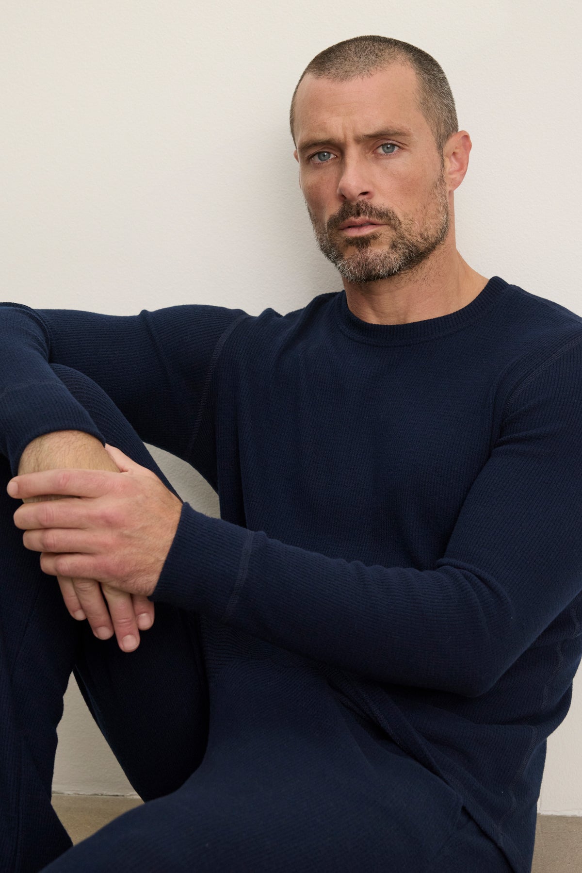 A man with short hair and a beard wears the TOBIAS CREW by Velvet by Graham & Spencer—dark blue classic thermal top and matching pants—as he sits against a light-colored wall with one knee raised.