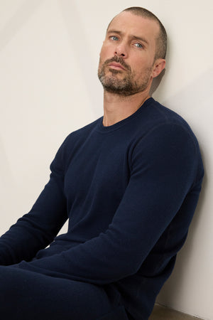 A man with short hair and a trimmed beard wears the Velvet by Graham & Spencer TOBIAS CREW, a cozy dark blue thermal sweater, as he sits against a light wall looking slightly upward.