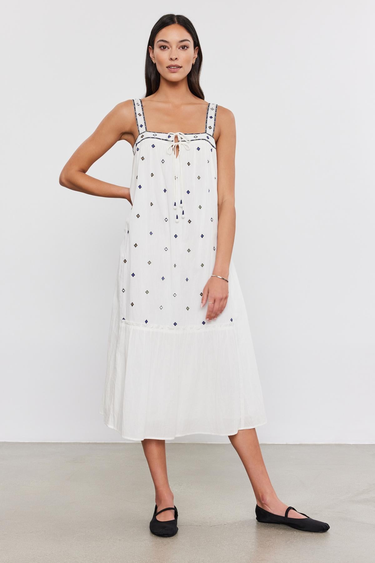   A woman in a white sleeveless Velvet by Graham & Spencer Riley dress with black polka dots and drawstring neckline, paired with black flat shoes, standing against a plain background. 