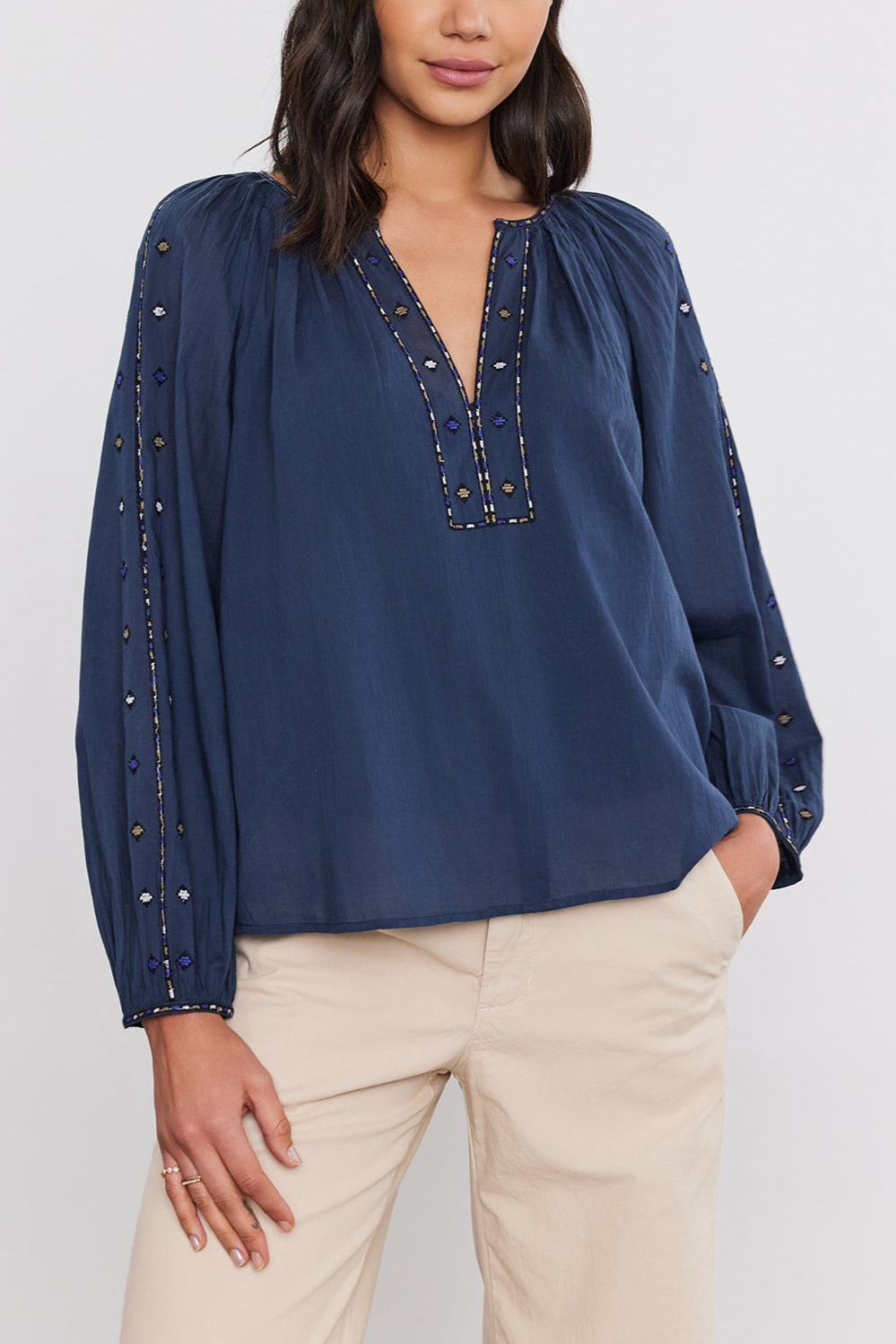 A woman with long dark hair wears a navy TINA TOP by Velvet by Graham & Spencer, featuring intricate crosshatch embroidery on the sleeves and neckline, paired with beige pants. She is standing against a plain white background.