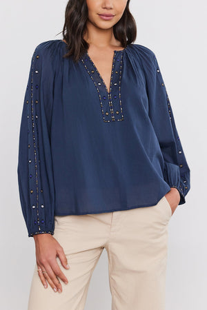 A woman with long dark hair wears a navy TINA TOP by Velvet by Graham & Spencer, featuring intricate crosshatch embroidery on the sleeves and neckline, paired with beige pants. She is standing against a plain white background.