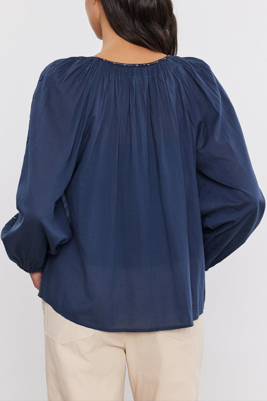 A person with long dark hair stands facing away, wearing a navy-blue, long-sleeved Velvet by Graham & Spencer TINA TOP with smocked detailing and light-colored pants against a plain background.-37452699697345
