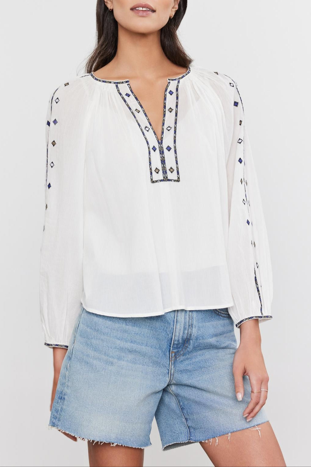 A woman wearing a "TINA TOP" by Velvet by Graham & Spencer with blue embroidery and denim shorts standing against a plain background.-36752975691969