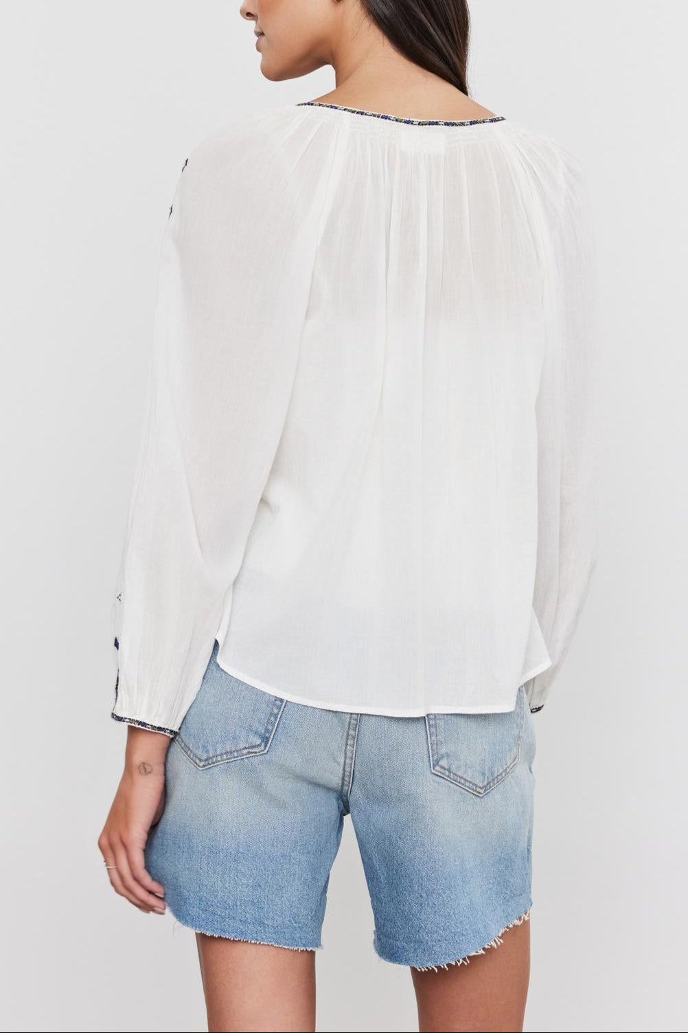 Woman in white TINA TOP blouse and denim shorts, viewed from the back, with a focus on the blouse's sheer texture and pleated details by Velvet by Graham & Spencer.