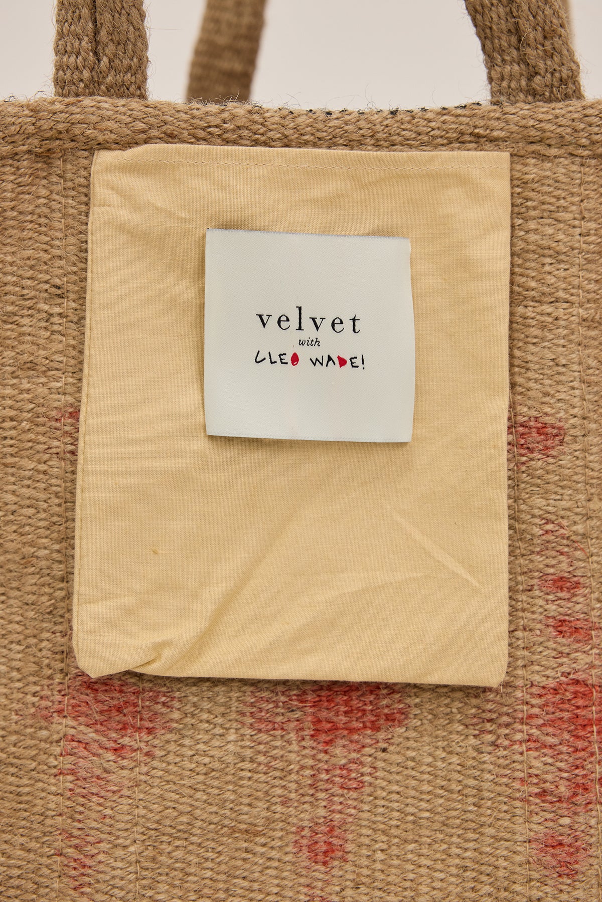 Close-up of the KADI BASKET BAG from Velvet by Graham & Spencer shows a woven jute design and a beige interior pocket. The pocket label reads "velvet with CLEO WADE!" in bold black and red text, with subtle red speckles on the bag's texture.-38864656335041