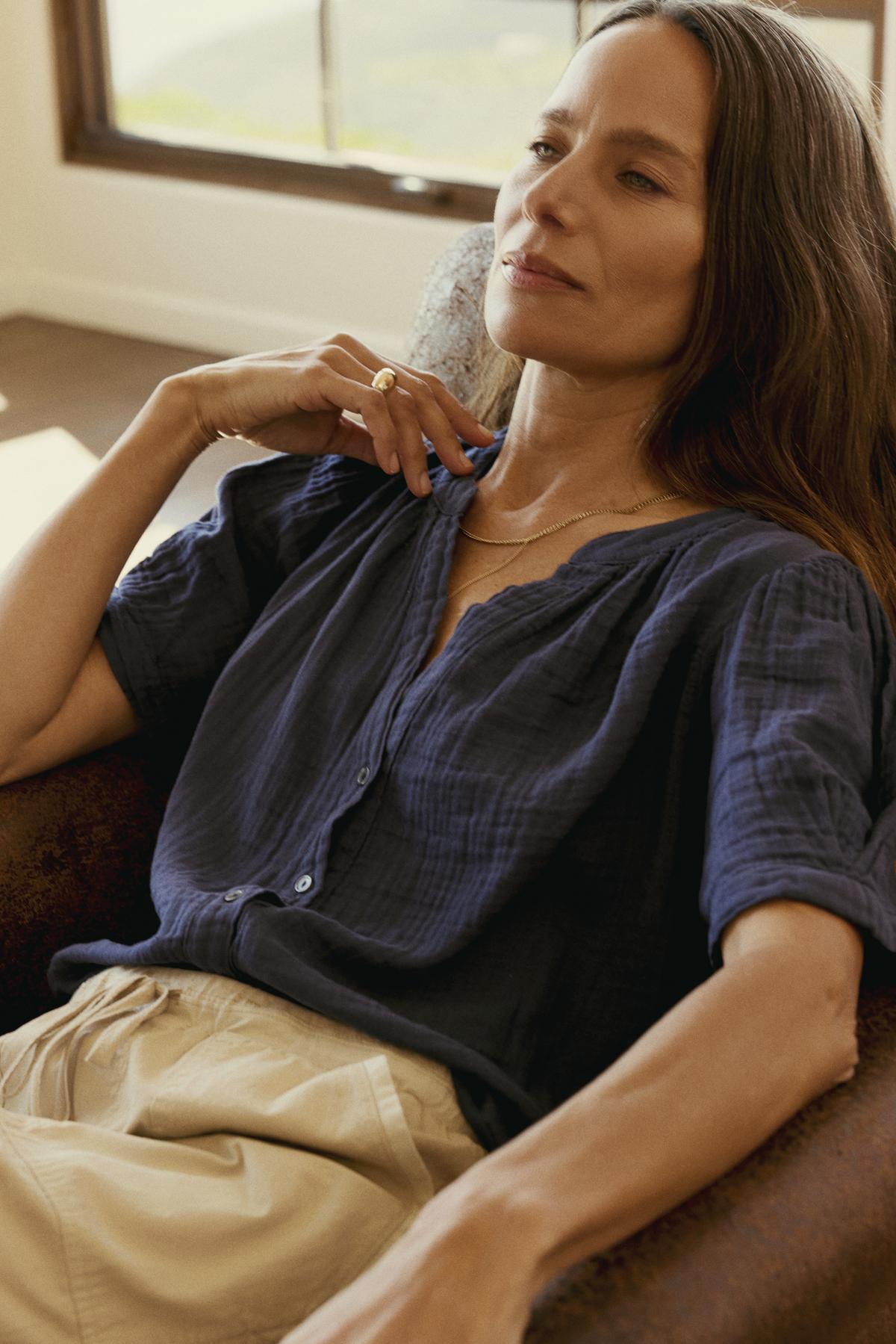 A woman with long hair is seated in a chair, wearing a dark blue DEANN COTTON GAUZE TOP by Velvet by Graham & Spencer and light-colored pants, with her left hand resting near her neck and a window in the background.-36443641970881