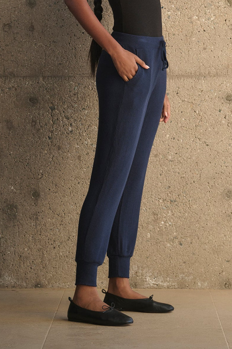 A woman with a long braid stands against a textured concrete wall, wearing a black tank top, the KOKO RIBBED JOGGERS from Velvet by Graham & Spencer featuring a drawstring waist in blue, and black loafers.-38282007969985