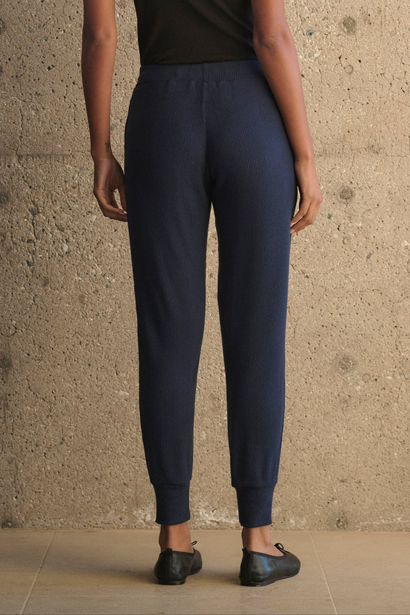 A person wearing a black tank top and Velvet by Graham & Spencer's KOKO RIBBED JOGGERS in navy, featuring a drawstring waist, stands with their back to the camera against a textured beige wall.-38282008002753