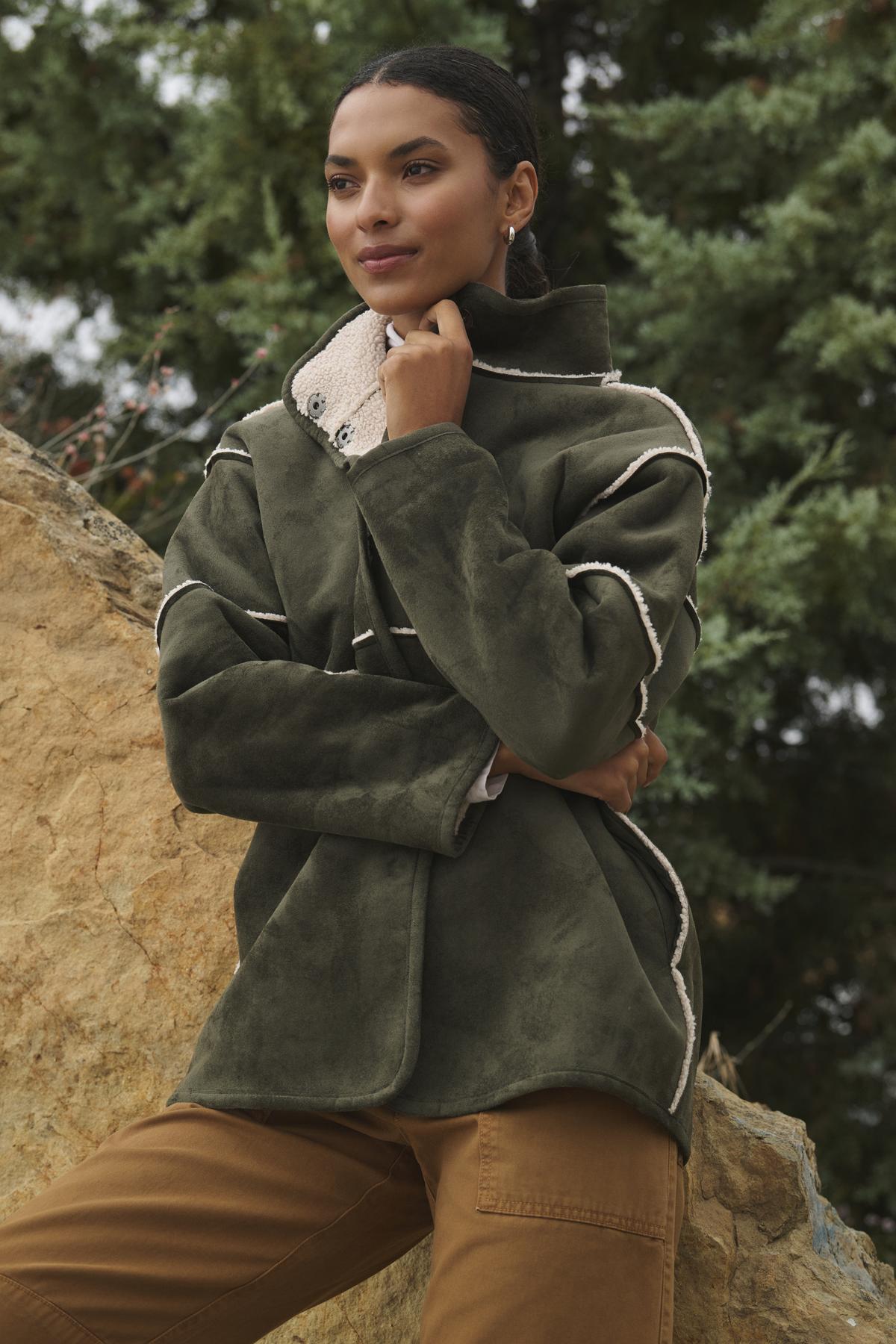 A person wearing the ALBANY LUXE SHERPA REVERSIBLE JACKET by Velvet by Graham & Spencer, featuring eco-friendly faux sherpa lining, stands outdoors, leaning against a rock with trees in the background.