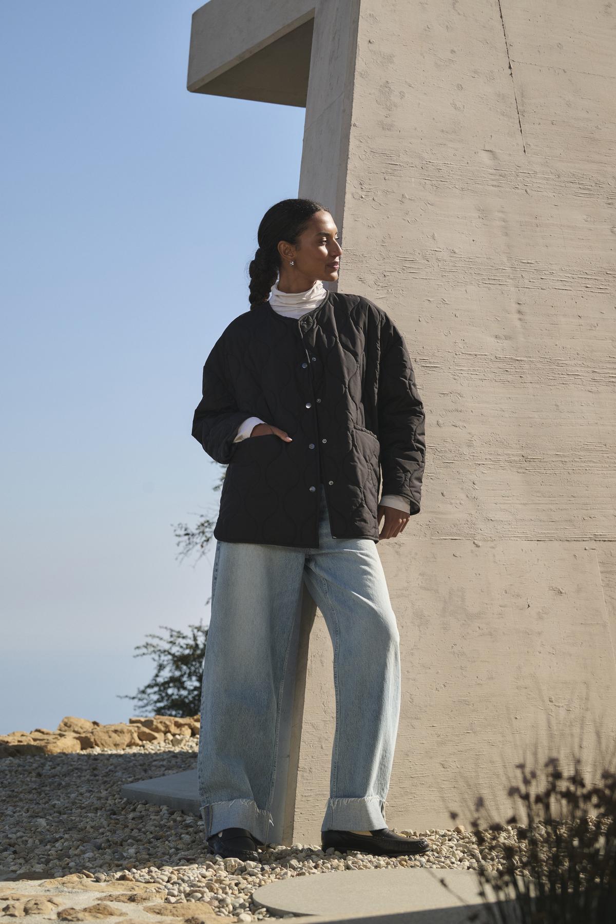 A person in a PAITYN QUILTED JACKET by Velvet by Graham & Spencer and jeans stands outdoors near a modern concrete structure, with a clear sky and rocky ground in the background.