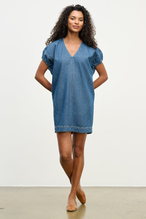 A woman models the AURORA DRESS by Velvet by Graham & Spencer—a short-sleeved, V-neck denim mini dress with a detachable belt—paired with beige flats, standing against a plain white background.