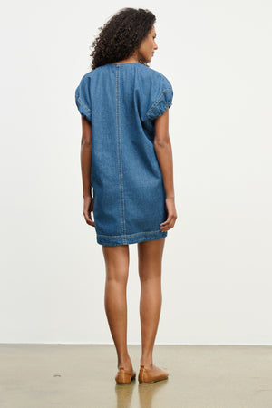 A woman faces away, modeling the AURORA DRESS by Velvet by Graham & Spencer—a blue denim mini dress with frayed edges and a detachable belt—paired with brown flats, set against a plain light background.