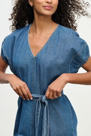A woman wearing the AURORA DRESS by Velvet by Graham & Spencer—a blue denim mini dress with short sleeves and a detachable belt—stands against a plain background.