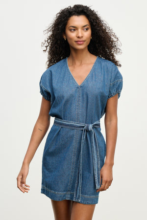 Model wearing the AURORA DRESS by Velvet by Graham & Spencer—a short-sleeved, V-neck denim mini dress with a detachable belt—stands against a plain light background.