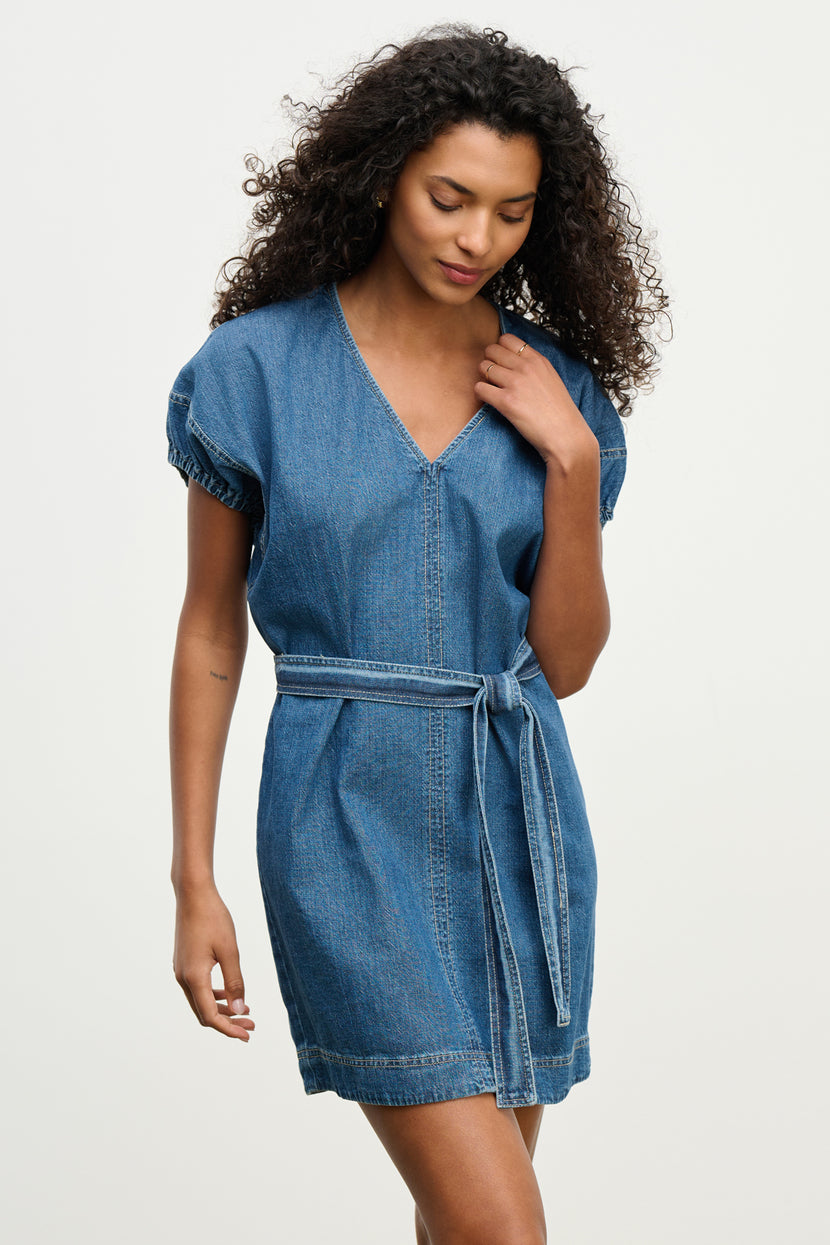 A woman models the AURORA DRESS by Velvet by Graham & Spencer, a short-sleeved denim mini dress with a V-neckline and detachable belt, set against a plain light background.