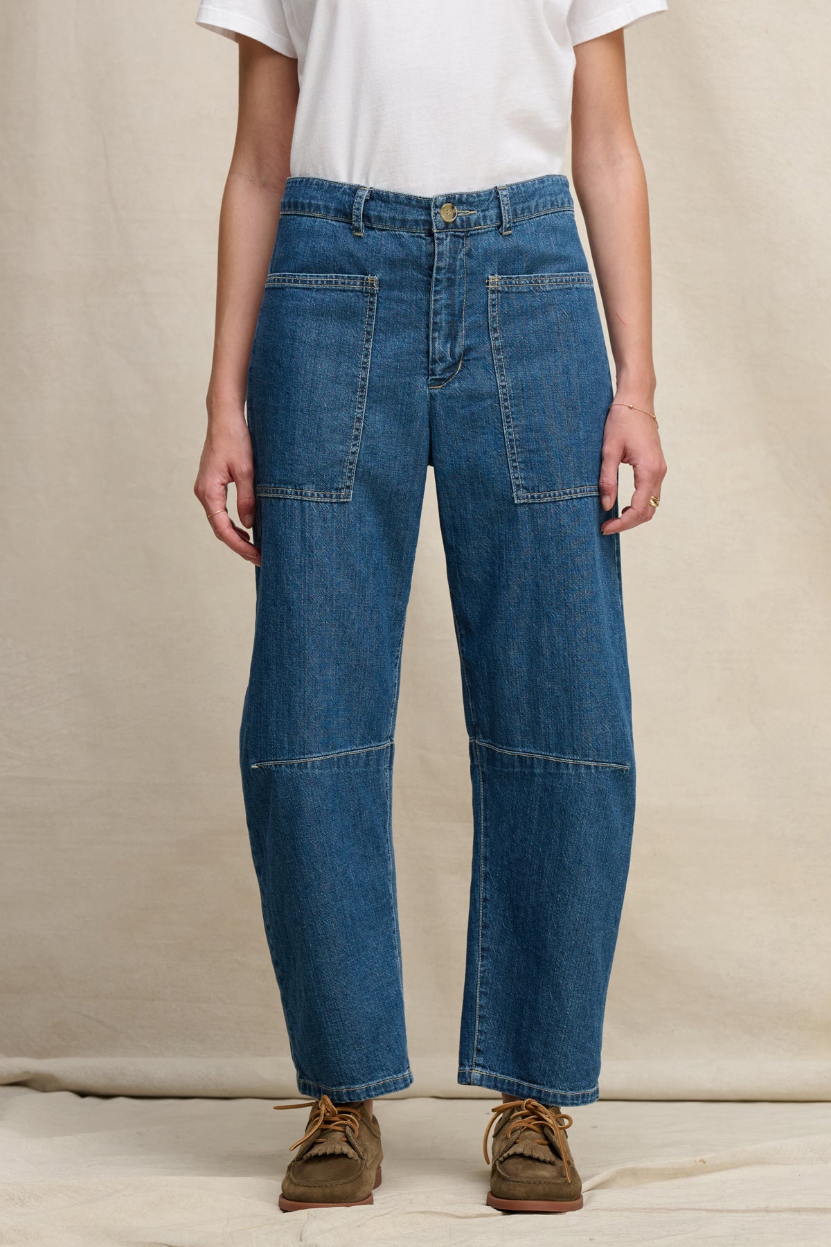 A person stands against a neutral fabric backdrop wearing a white t-shirt, brown shoes, and the FARAH DENIM PANT by Velvet by Graham & Spencer—blue high-waisted jeans with a curved leg silhouette and large patch pockets.