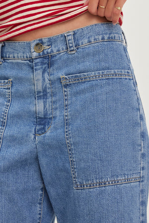 A person is wearing the FARAH DENIM PANT by Velvet by Graham & Spencer, featuring blue denim with large front pockets and a button closure. A red and white striped top is partially visible.
