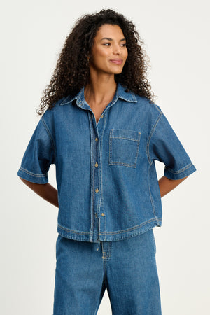 A person with curly hair wearing the KADDY DENIM TOP by Velvet by Graham & Spencer, a relaxed fit, short-sleeved denim top paired with matching denim pants, stands with hands behind their back.
