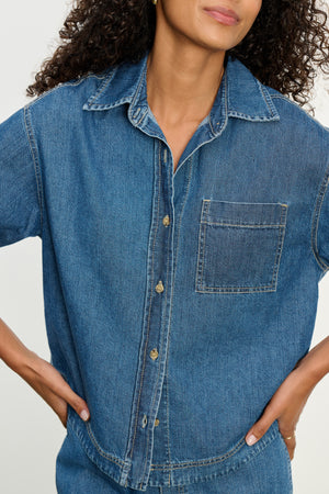 A person wearing the KADDY DENIM TOP by Velvet by Graham & Spencer, a relaxed fit, short-sleeve denim shirt with a button-front and chest pocket, stands with hands on hips.