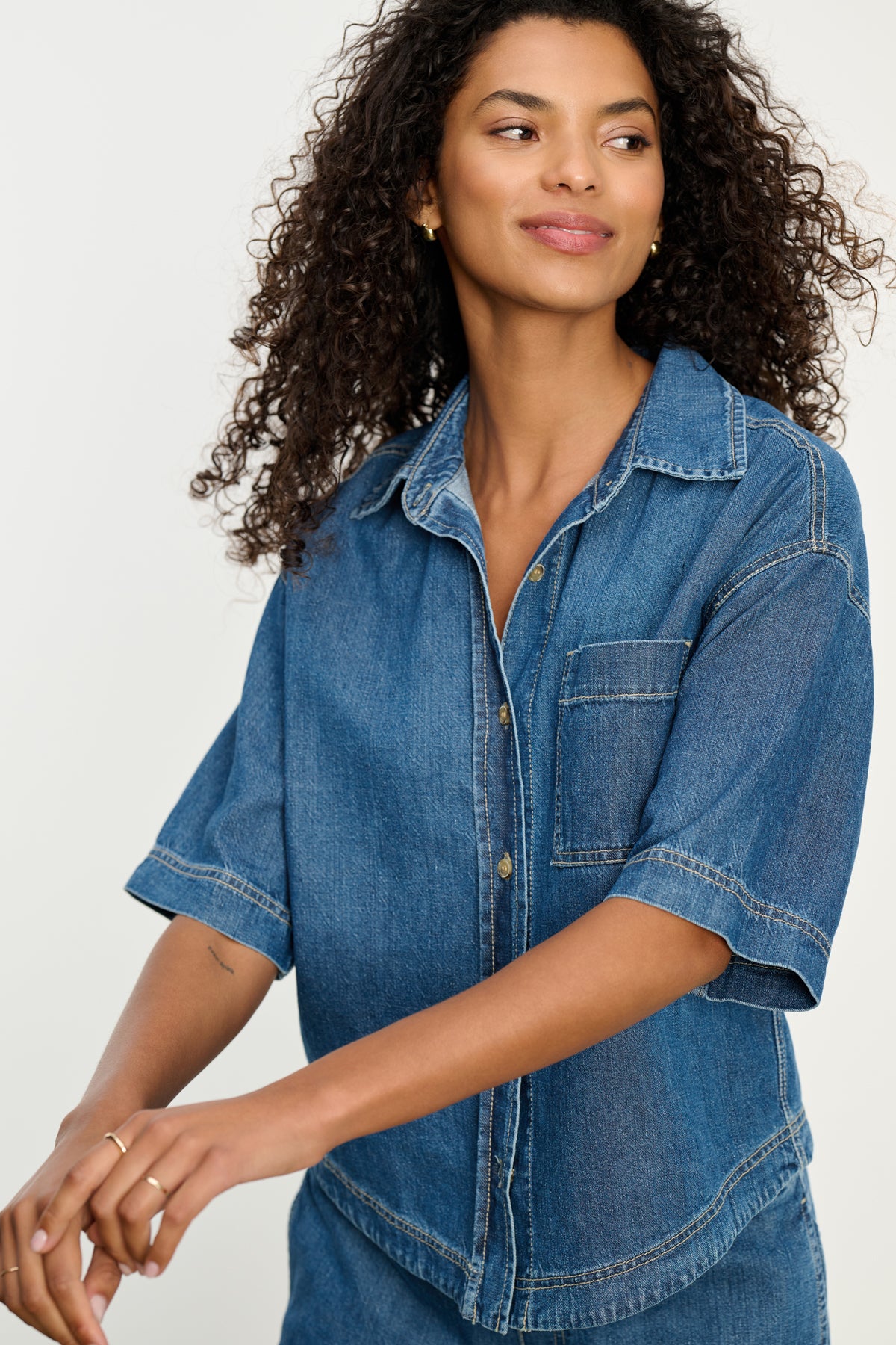 A woman with curly hair models the KADDY DENIM TOP by Velvet by Graham & Spencer and matching pants in a relaxed fit, standing against a plain background.