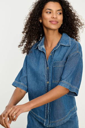 A woman with curly hair models the KADDY DENIM TOP by Velvet by Graham & Spencer and matching pants in a relaxed fit, standing against a plain background.