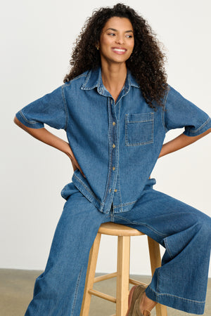 A woman with curly hair sits on a wooden stool, wearing the Velvet by Graham & Spencer KADDY DENIM TOP with a chest pocket and matching wide-leg denim pants.