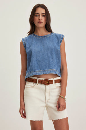 A person wearing the sleeveless LOREN DENIM TOP by Velvet by Graham & Spencer, cream-colored shorts, and a brown braided belt stands against a plain background.