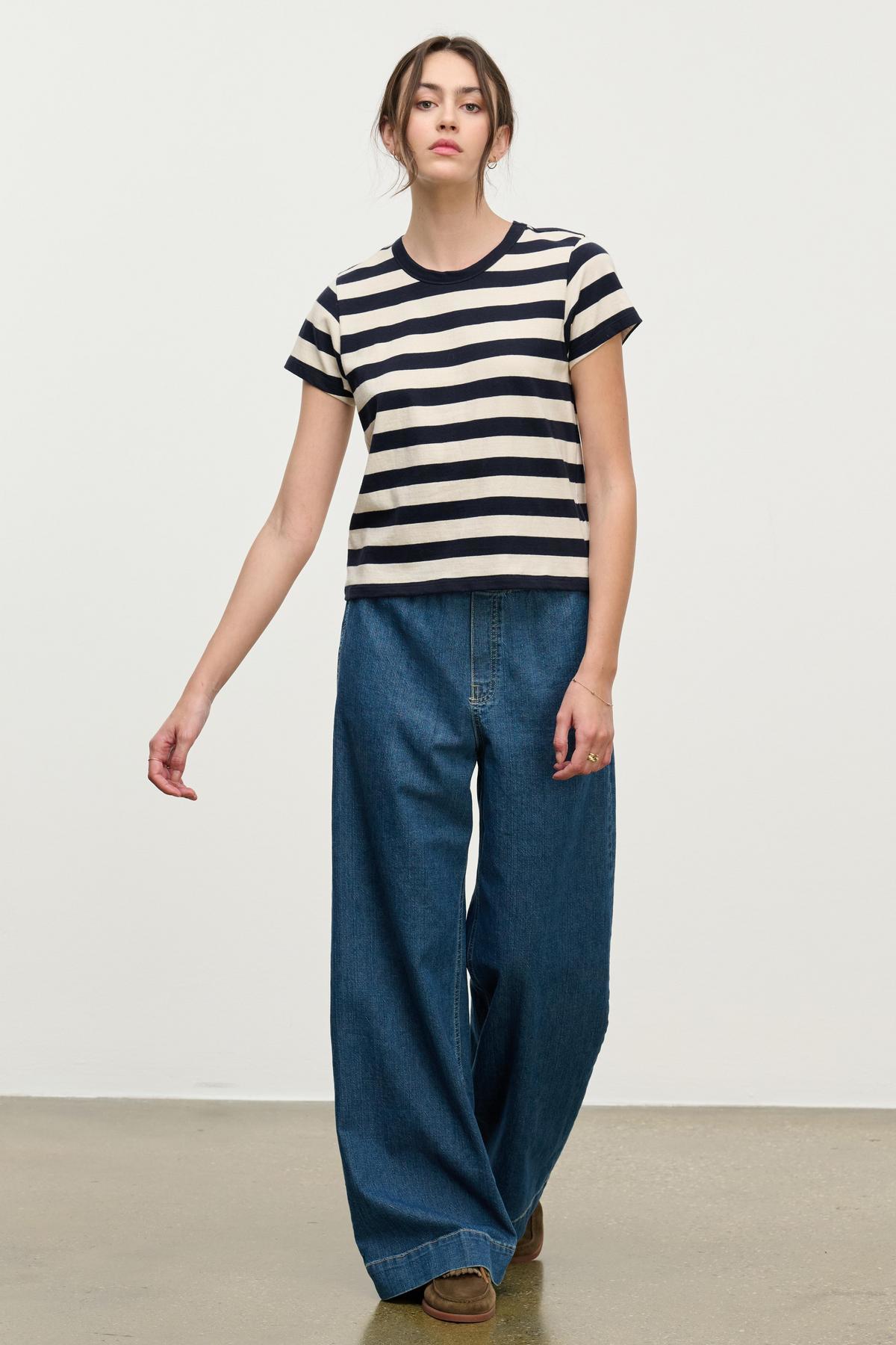 A person stands against a plain background wearing a short-sleeved, black and white striped shirt, brown shoes, and Velvet by Graham & Spencer’s MIRA DENIM PANT featuring wide legs and a drawstring waist.-39742352031937
