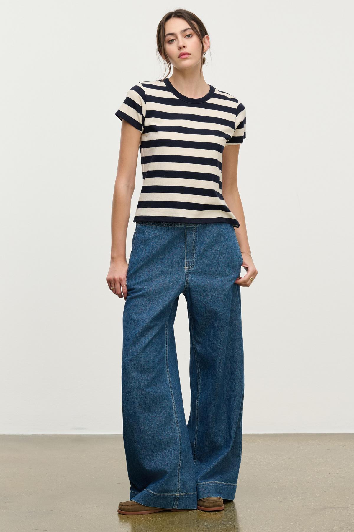 A person stands against a plain background wearing a black and white striped T-shirt, brown shoes, and the MIRA DENIM PANT by Velvet by Graham & Spencer—wide-leg denim pants featuring a drawstring waist.