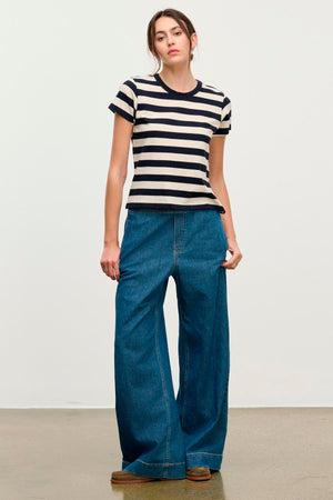 A person stands against a plain background wearing a black and white striped T-shirt, brown shoes, and the MIRA DENIM PANT by Velvet by Graham & Spencer—wide-leg denim pants featuring a drawstring waist.