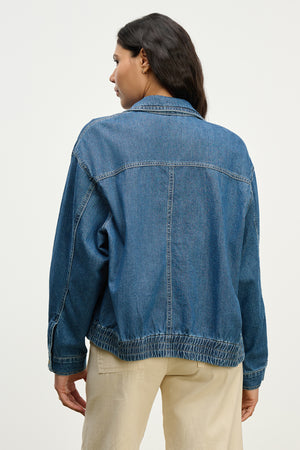 A woman is seen from the back in the PORTER DENIM ZIP-UP JACKET by Velvet by Graham & Spencer, featuring an elastic hem, paired with beige pants against a plain light background.