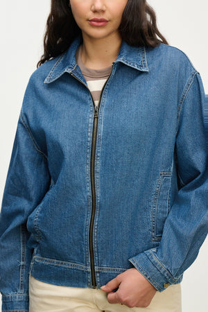 A person wearing the PORTER DENIM ZIP-UP JACKET by Velvet by Graham & Spencer over a striped shirt and light pants, holding the jacket's elastic hem.