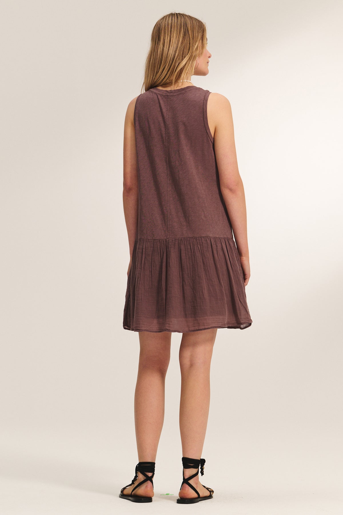 A woman faces away, wearing the DENNA DRESS by Velvet by Graham & Spencer—a sleeveless, loose brown dress with a gathered skirt—paired with black sandals against a plain background.-39173058625729