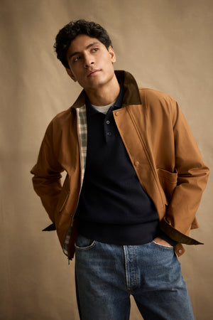 A man stands with his hands in his pockets against a plain beige background, wearing the DESREN JACKET by Velvet by Graham & Spencer—a brown cotton canvas jacket with a corduroy collar—over a dark sweater and blue jeans.