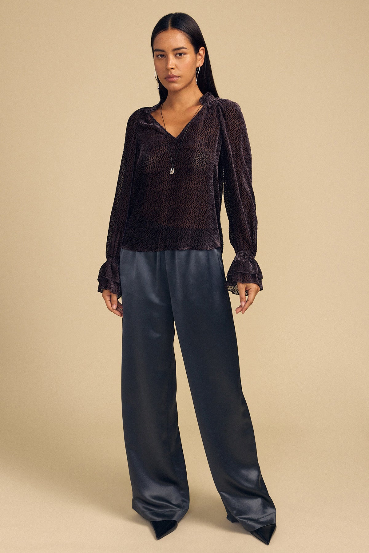 A woman wearing an EMERSON DEVORE DOT BLOUSE in dark silk with a delicate dot pattern, ruffled collar and cuffs, and wide-leg trousers.-46488255463617