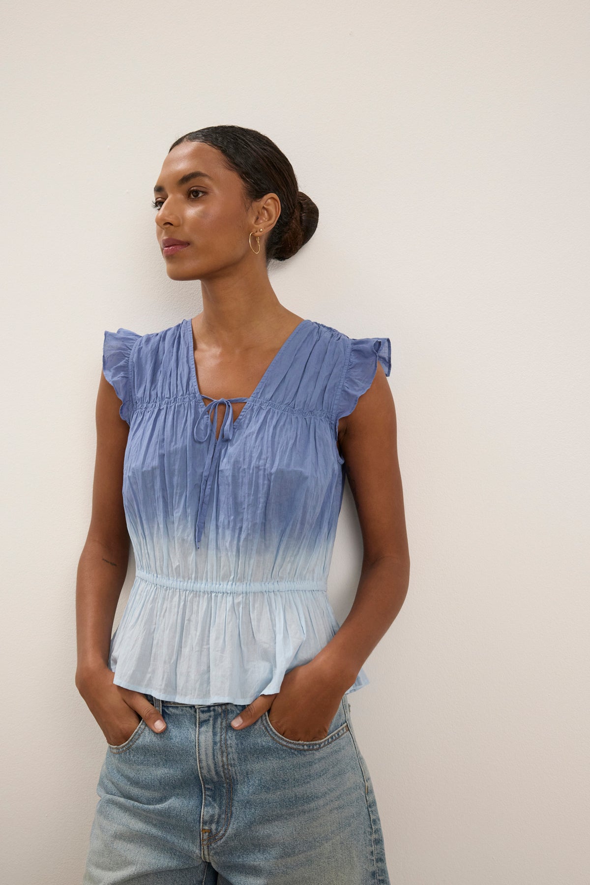 A woman stands against a beige wall, wearing the KAIA BLOUSE by Velvet by Graham & Spencer—blue dip-dye with ruffled shoulders, V-neckline, crinkle voile texture—paired with light blue jeans and hands in pockets.-39111853080769
