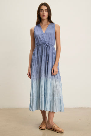 A woman stands indoors wearing the Velvet by Graham & Spencer MARILYN DRESS, a sleeveless, wrap-front V-neck blue dip-dye maxi with a detachable belt and tan sandals. She faces forward, arms relaxed at her sides.