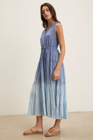 A woman stands indoors in the Velvet by Graham & Spencer MARILYN DRESS—a sleeveless, blue ombre tiered maxi with a tie waist—paired with flat beige sandals, facing sideways against a plain background.
