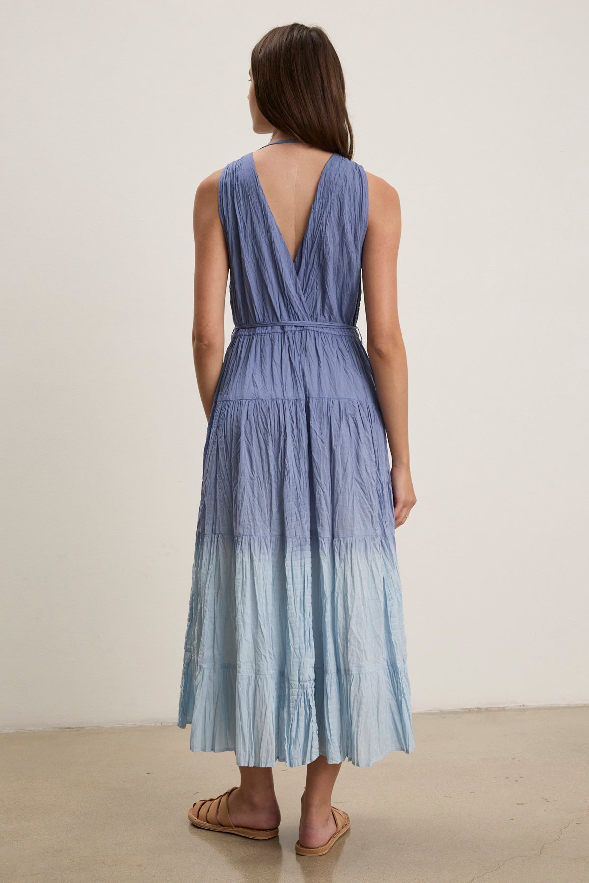 A woman stands indoors facing away, wearing the sleeveless MARILYN DRESS by Velvet by Graham & Spencer, a dip-dye crinkle voile maxi dress with a V-back, paired with flat sandals.