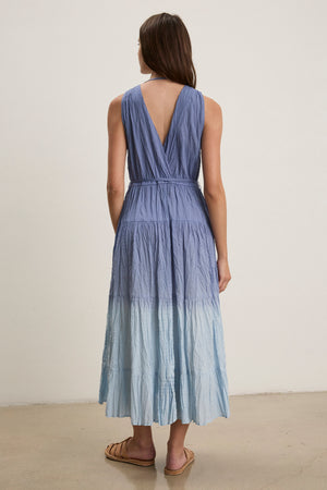 A woman stands indoors facing away, wearing the sleeveless MARILYN DRESS by Velvet by Graham & Spencer, a dip-dye crinkle voile maxi dress with a V-back, paired with flat sandals.