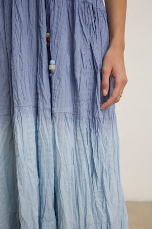 A person in the MARILYN DRESS by Velvet by Graham & Spencer, featuring a feminine fit and light blue dip-dye crinkle voile with beaded drawstrings, stands with one hand visible, adorned with a simple gold ring.