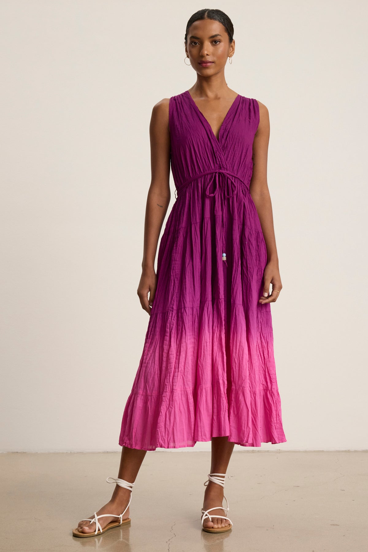 A woman stands indoors wearing the MARILYN DRESS by Velvet by Graham & Spencer, a sleeveless wrap-front V-neck dip-dye crinkle voile midi dress in purple-to-pink ombre, paired with white strappy sandals.