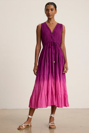 A woman stands indoors wearing the MARILYN DRESS by Velvet by Graham & Spencer, a sleeveless wrap-front V-neck dip-dye crinkle voile midi dress in purple-to-pink ombre, paired with white strappy sandals.