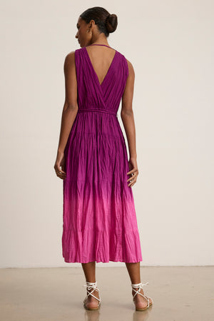 A woman, hair in a bun and wearing sandals, faces away in the Velvet by Graham & Spencer MARILYN DRESS—an ankle-length, sleeveless crinkle voile dress with a purple-to-pink dip-dye gradient, V-back, and detachable belt.