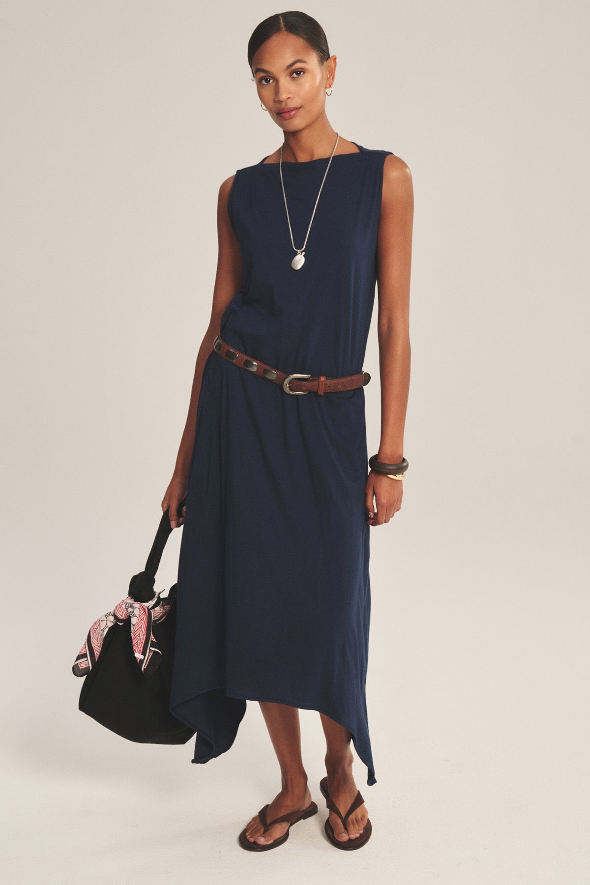Woman wearing the DREE DRESS by Velvet by Graham & Spencer, a sleeveless navy style with a handkerchief hem and brown belt, paired with sandals, jewelry, and a black bag adorned with a pink scarf.-41008618373313