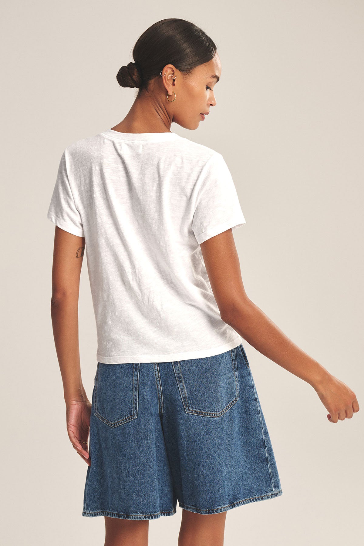 A woman stands facing away, wearing the ELISHA TEE by Velvet by Graham & Spencer—a white cotton slub V-neck tee paired with blue denim shorts. Her hair is tied in a bun, highlighting this effortless wardrobe staple.-41629729292481