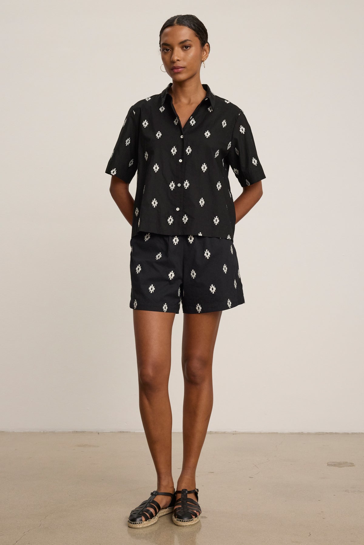 A person stands in front of a plain background wearing Velvet by Graham & Spencer's DAPHNE Embroidered Cotton Voile Shorts with an elastic waistband and a white geometric pattern, paired with a matching black shirt and black sandals.-39488863764673