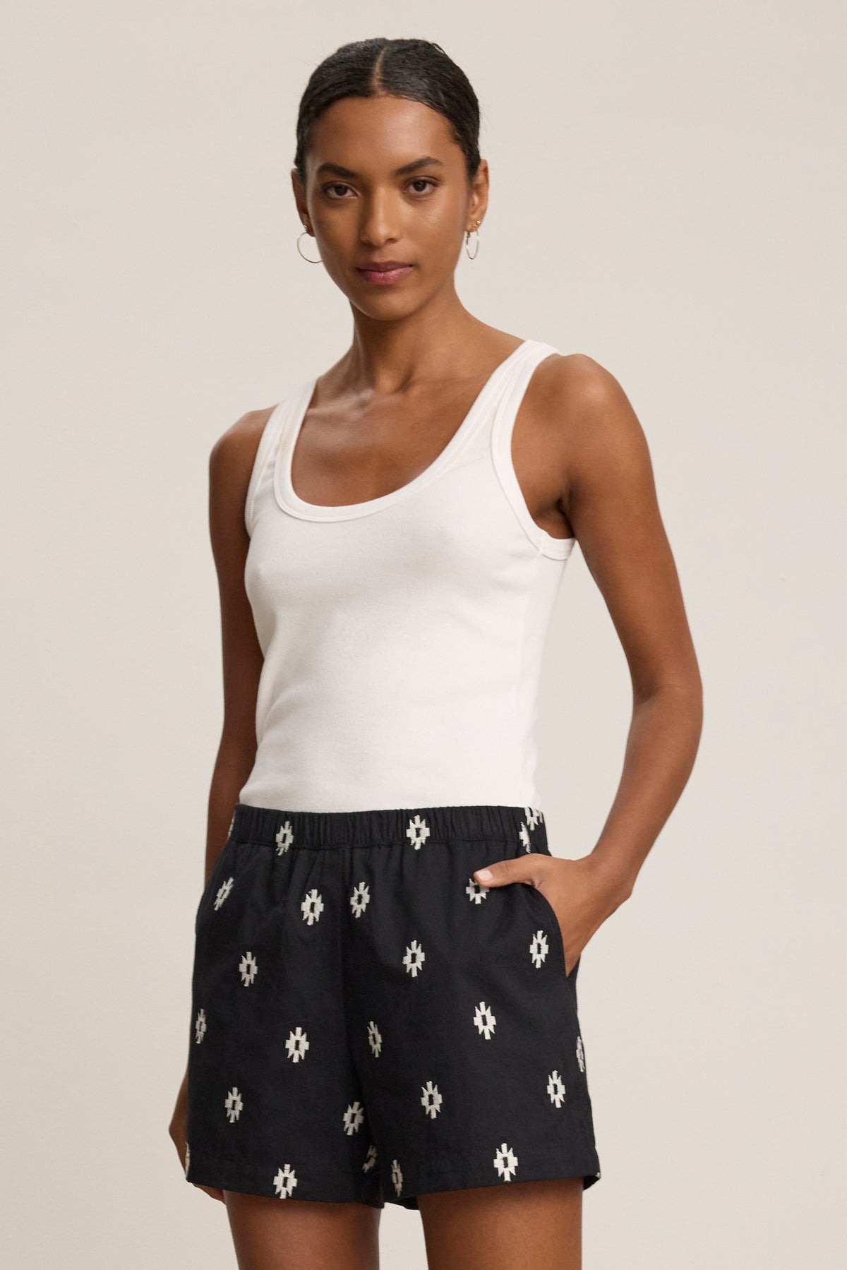 A woman poses against a plain background in a white tank and Velvet by Graham & Spencer's DAPHNE EMBROIDERED COTTON VOILE SHORT, featuring a white geometric pattern, elastic waistband, and one hand in her pocket.