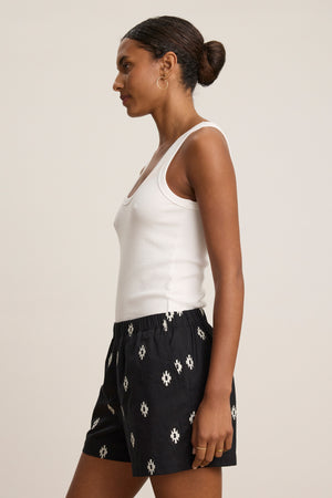 A woman in profile wears a white sleeveless top and Velvet by Graham & Spencer's DAPHNE EMBROIDERED COTTON VOILE SHORT, featuring a black breathable fabric with a white geometric pattern and an elastic waistband, against a plain light background.